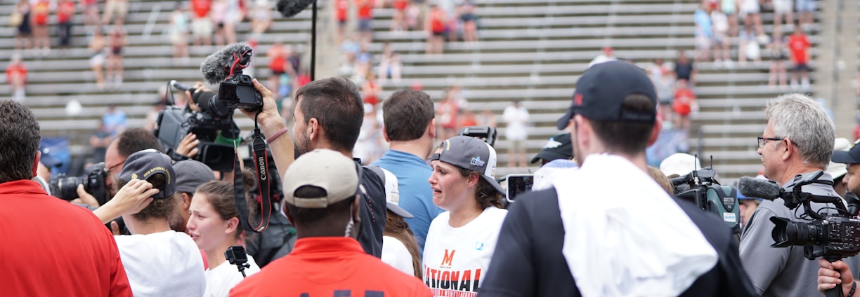 A lively gathering in an outdoor sports venue with a group of people, some wearing matching shirts labeled 'National Champions.' Several individuals are holding cameras and microphones, indicating a media presence. The background shows blurred spectators sitting on bleachers.